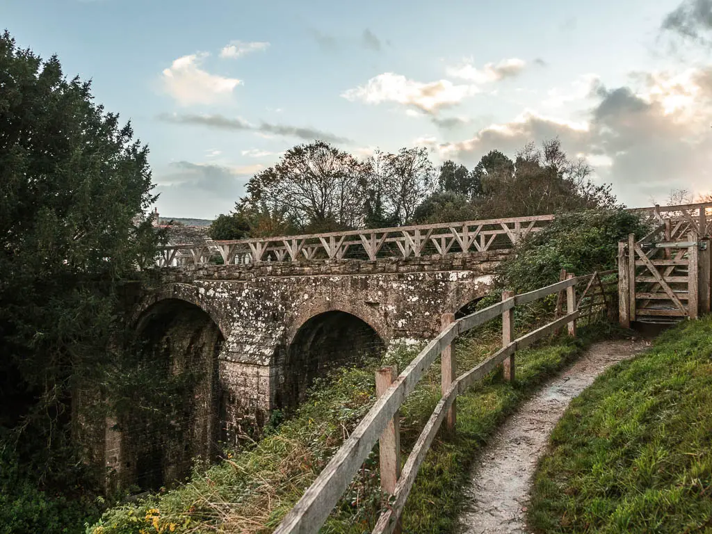 A narrow path on the right, lined with a wooden fence, and a sort bridge ahead as you enter Corfe at the end of the walk from Wareham.