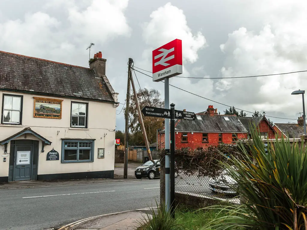 A road with a Wareham railway sign pin red next to it, at the start of the walk. There is a white walled pub on the other side of the road.