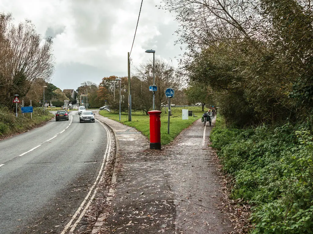 A road on the left, and walking path on the right. The road and path are split by the green ahead. There is a red post box in between the road and path.