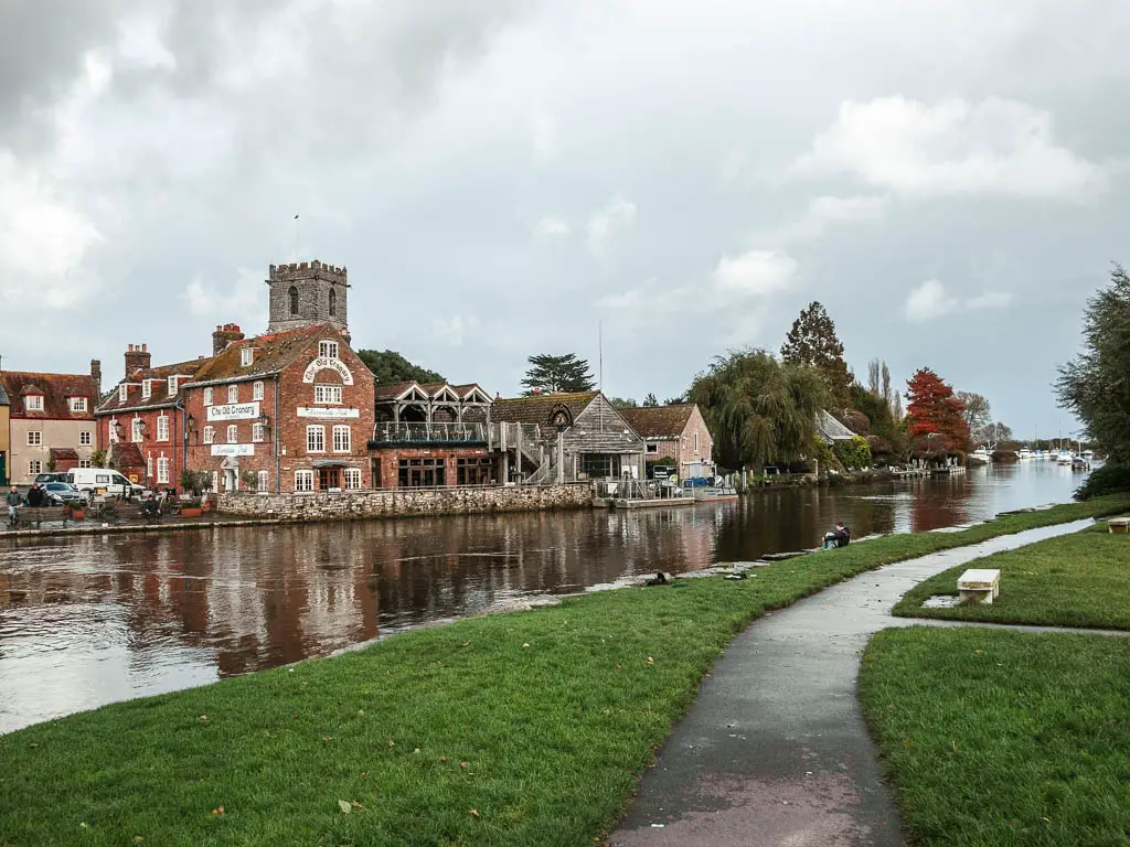 A walking path leading straight, on the right, and the river Frome on the left, at the start of the walk out of Wareham. There are old buildings on the other side of the river.