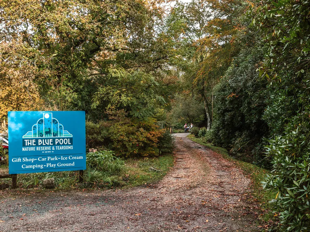 A widę gravel path leading ahead on the right, and a big blue sign on the left saying 'the blue pool'. The path is lined with green leafy bushes and trees.