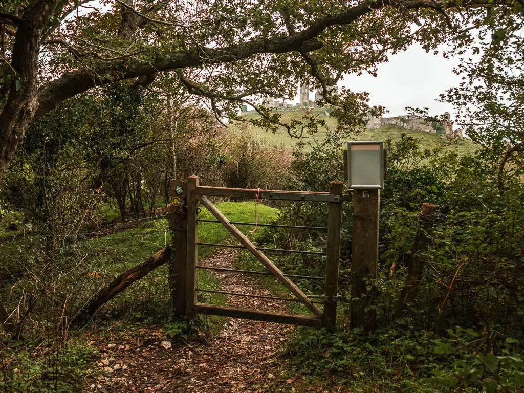 A wooden gate across the narrow dirt trail, surrounded with bushes, and a tree hanging over it.