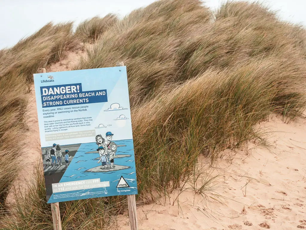 A danger warning sign in blue in front of the tall grass on the sand dune.