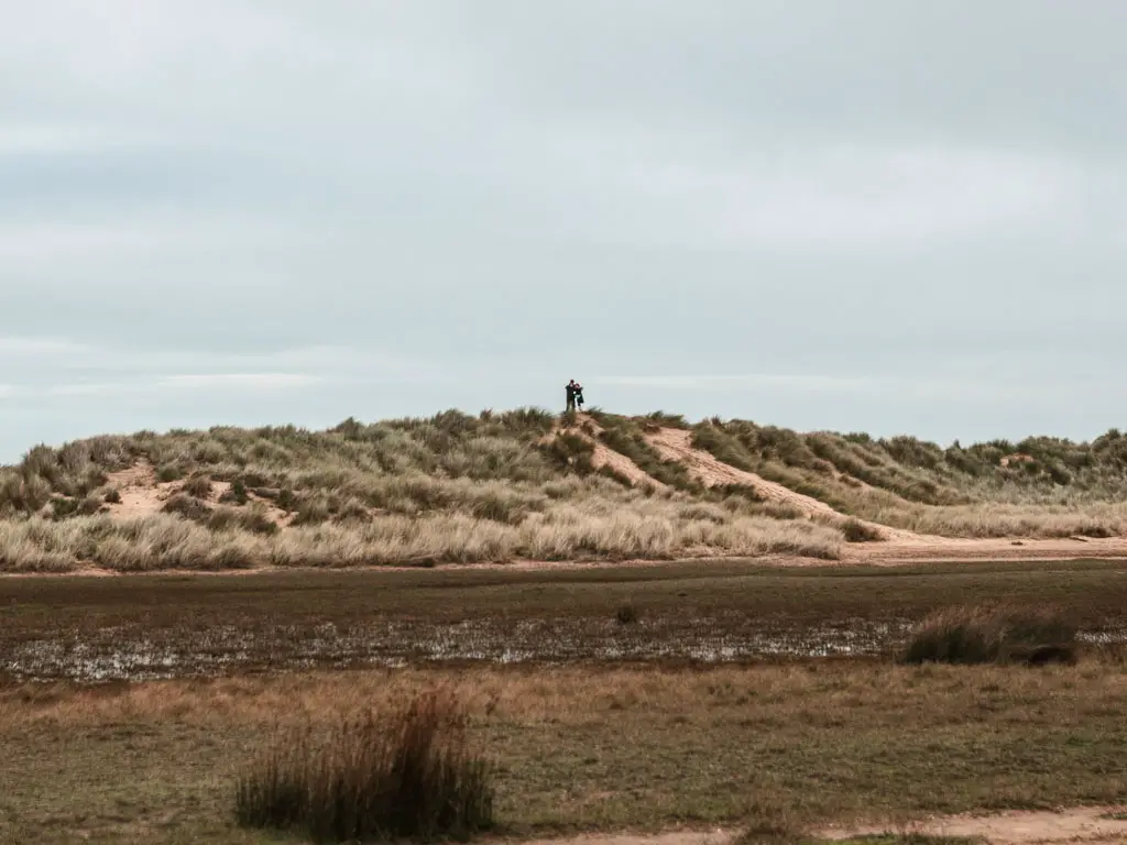 Looking across the boggy ground to sand dunes covered in grass on the other side. There are two people standing on top of the sand dune.