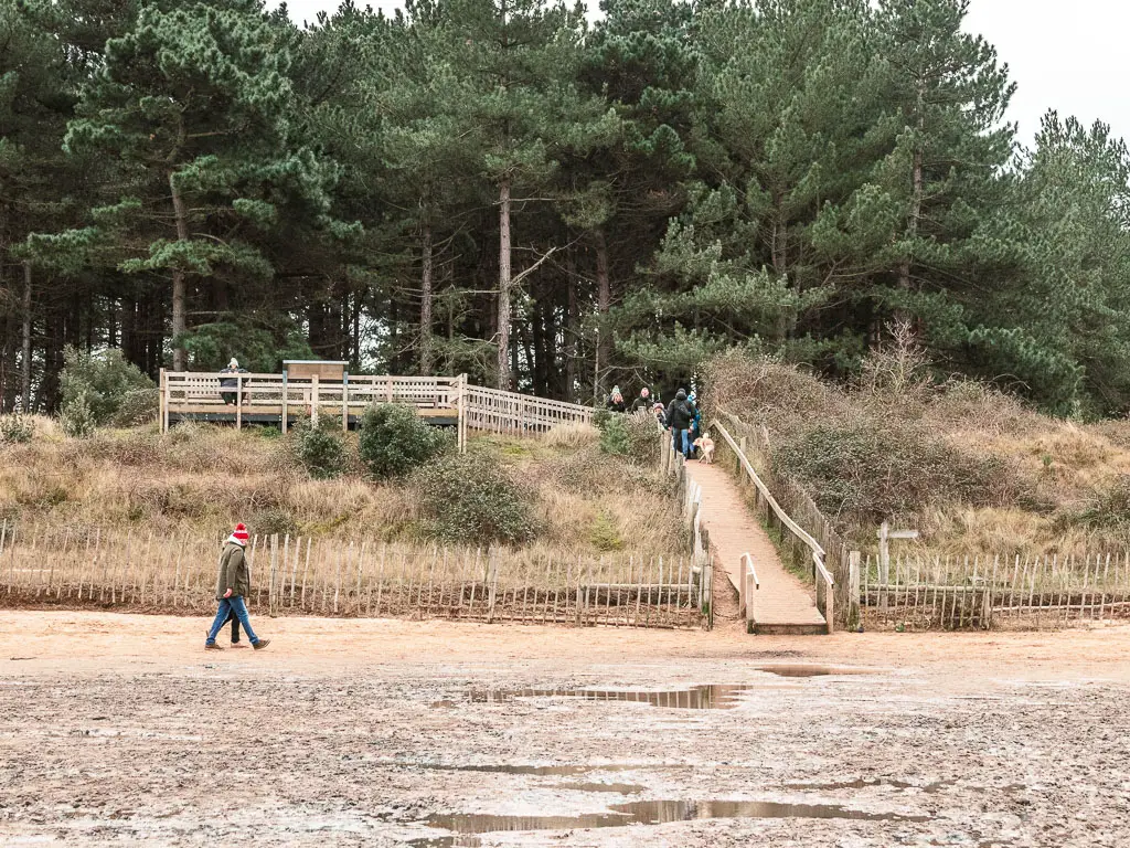 Looking across the boggy sand to a walkway leading up off the beach to the woods. There are people walking along the beach and up the walkway.