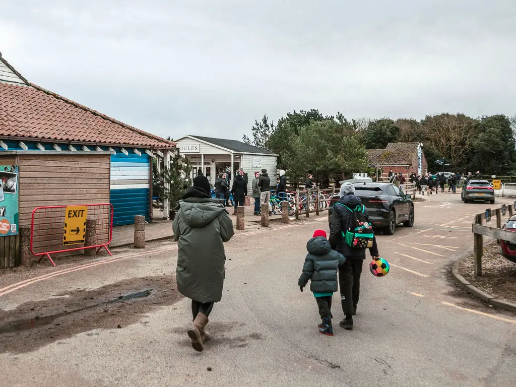 Lots of people walking along the road through the car park, with refreshment huts ahead.