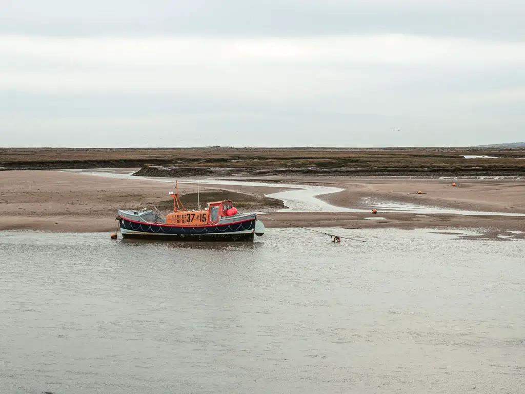 A small boat sitting on the low river next to the sandy bank, part way through the walk between Burnham Overy Staithe and Blakeney. The boat is black underneath, and has an orange top.