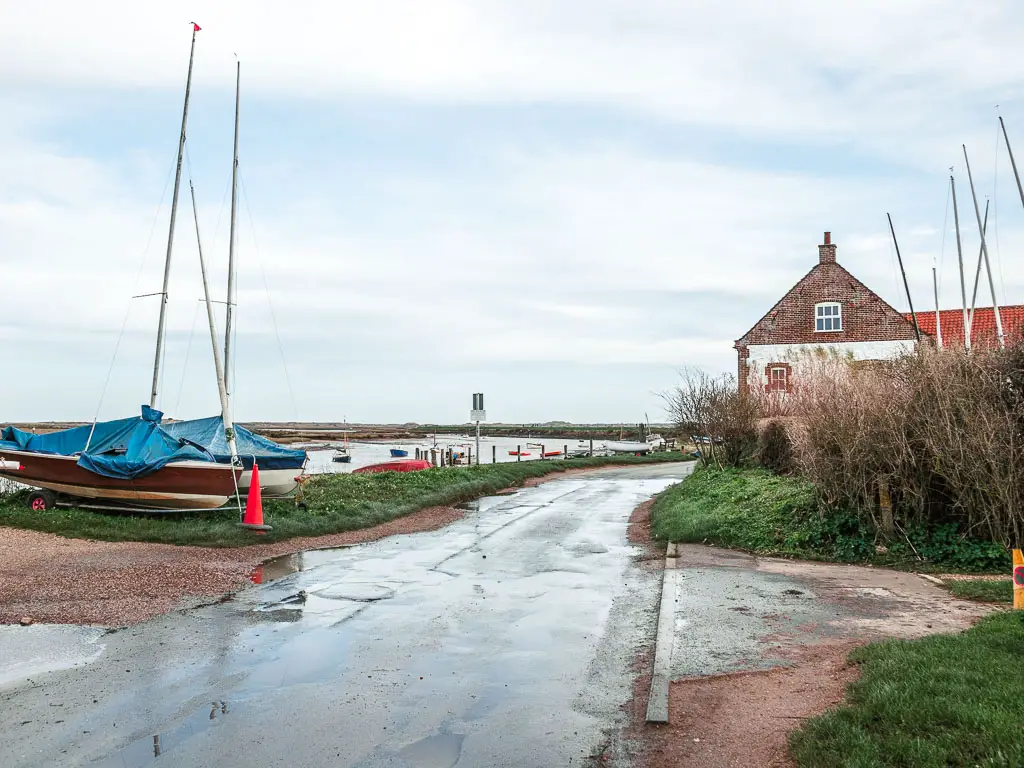 A road with two sailboats on the green bank on the left side. There is a brick building on the right behind some bushes, and the river visible ahead to the left.