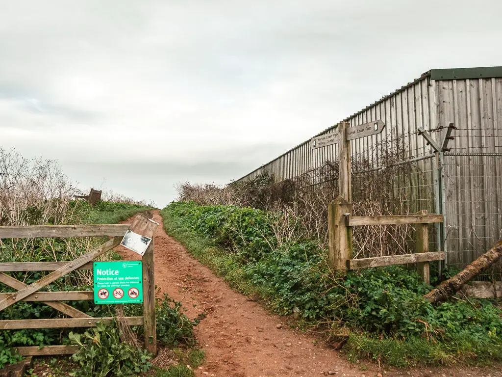 A dirt walking trail leading through a wooden fence, and lined with green grass and bushes. 