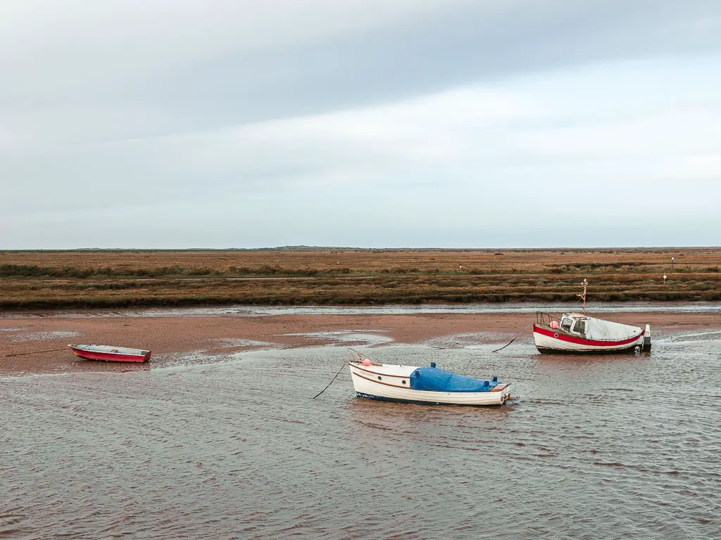 Three small boats in the river, at the start of the walk from Burnham Overy Staithe to Blakeney. The river is low and there are marshes on the other side. 