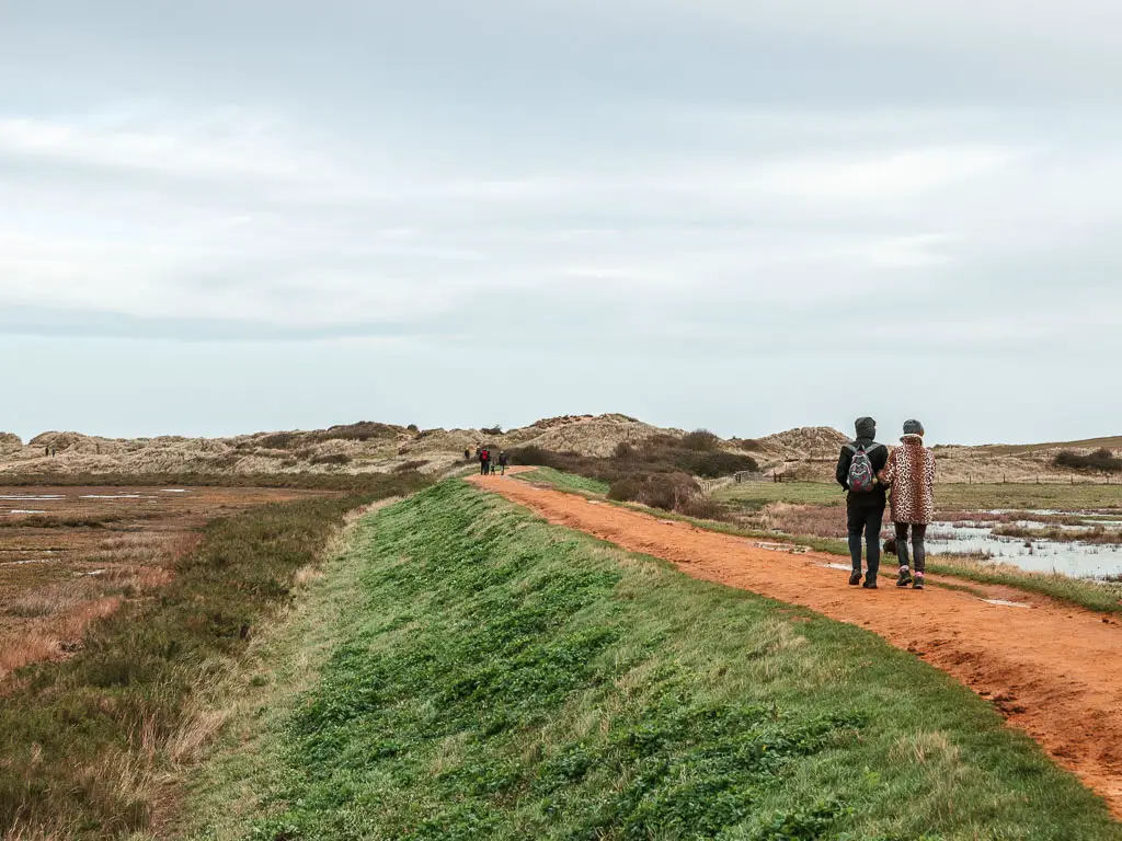 People walking along the dirt trail ridge, towards sand dunes ahead. There is green grass down the left side of the ridge, with marshes to the left. 