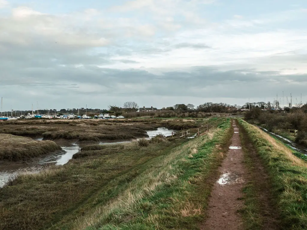 A dirt ridge walking trail, with grass sides. There is marshland to the left and boats in the distance to the left.