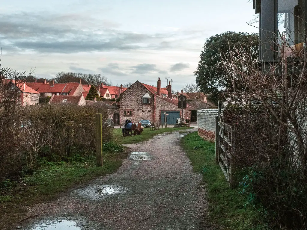 A gravel path leading through a gate and bushes to the stone walled buildings of Blakaney ahead.