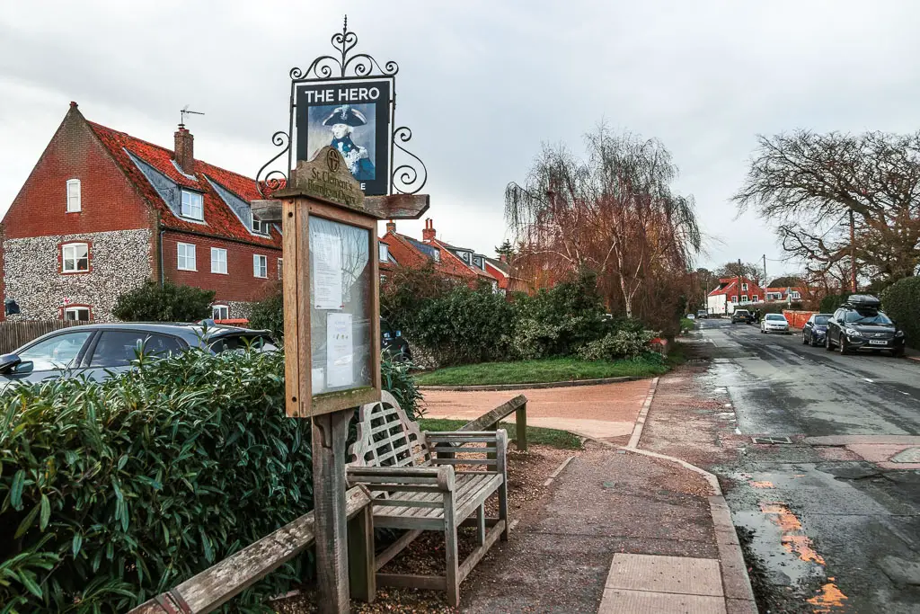 A bench and sign for the Hero Pub on the pavement, with the road on the right. There are houses on the left side of the road, and cars parked on the right side.