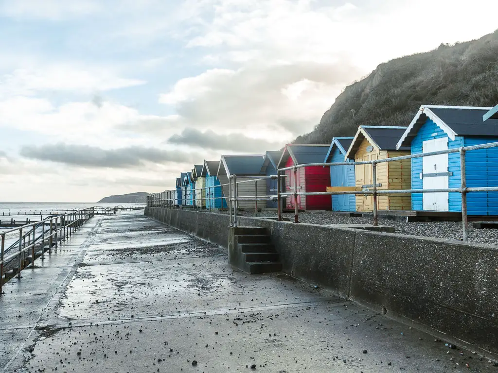 A grey concrete walkway, with an upper level on the right with a row of colourful beach huts, when walking from Cromer to Mundesley.