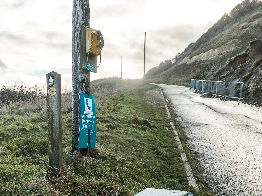 A road leading uphill on the right, and strip of grass lining it on the left. There is a big wooden pole with a yellow telephone on the left, as well as a smaller wooden post with a white acorn sign.