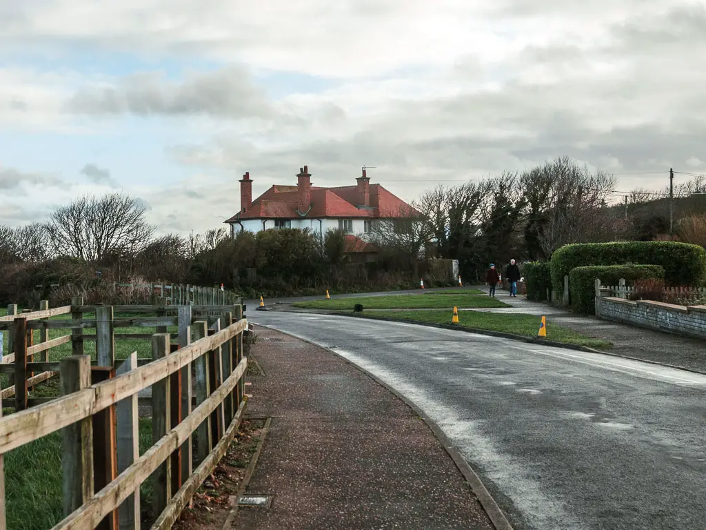 A road curving ahead to the left, with a pavement lining the left side, and green on the other. There is a big white walled house with a red roof on the other side of the road, partially hidden by bushes.