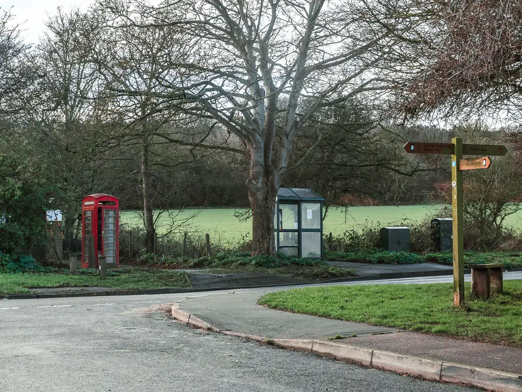 A road junction, with a red telephone box on the other side. There is a wooden trail sign on the corner of the junction, pointing, left, right, and back.