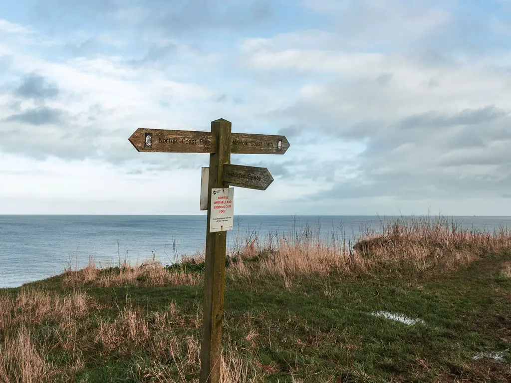 A wooden trail sign on the grass clifftop, with the blue sea ahead. The sign points ahead, back, and right. 