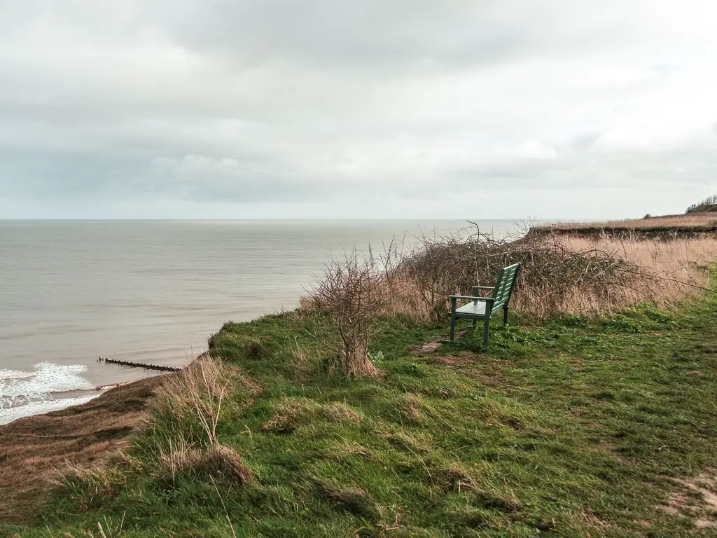 A small green bench on the grass clifftop, with a view to the sea below to the left. The bench is facing the sea.