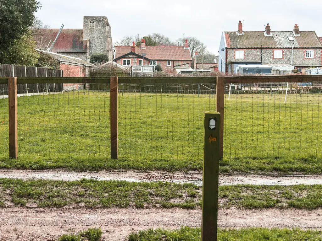 A wooden stump signpost with a white acorn and arrow pointing left. There is a wire fence behind it, leading onto a grass field and houses on the other side.