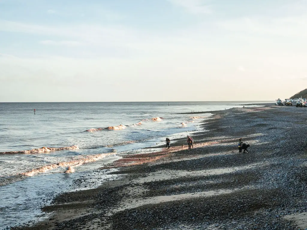 Looking down and along a black shingle beach in Cromer, as it meets the wavy sea on the left, at the start of the walk towards Mundesley. There are a few people on the beach.