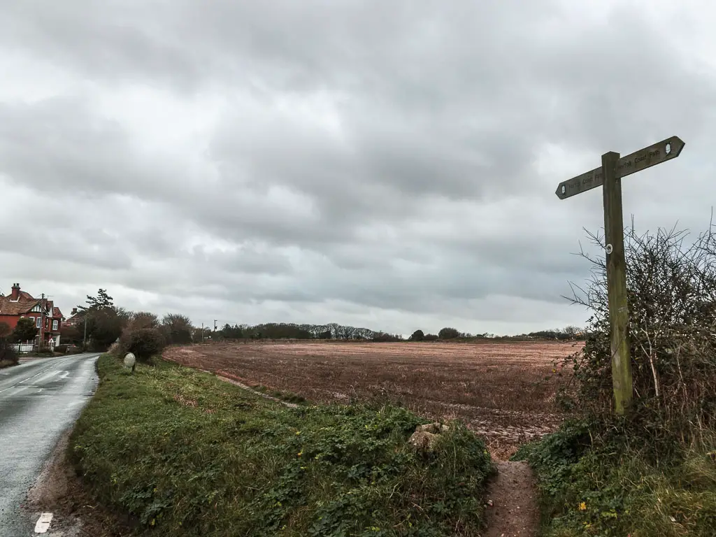 A wooden trail sign pointing ahead along the edge of a brown crop field. There is a road on the left side of the field.