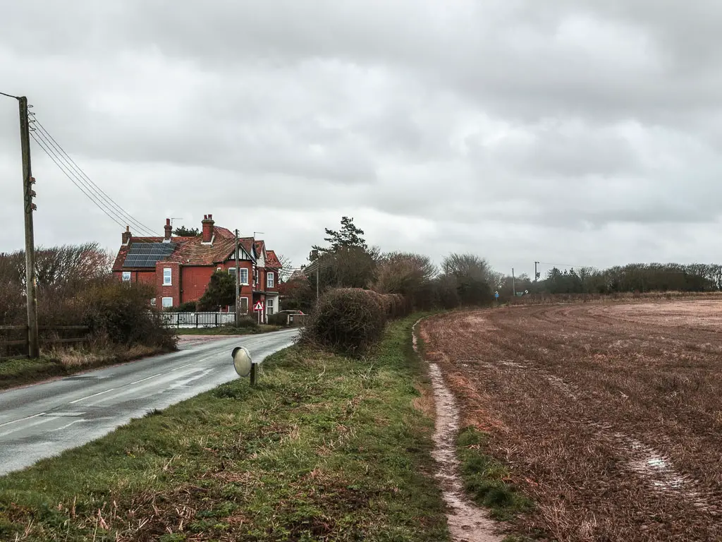 A narrow trail with a brown crop field to the right, and the road to the left. There is a house on the left side of the road ahead.