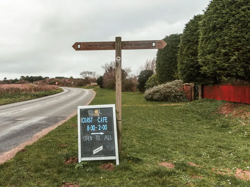 A wooden trail signposts with a cafe blackboard sign below it, sitting on the grass, with the road to the left. The arrows on the sign point left and right.