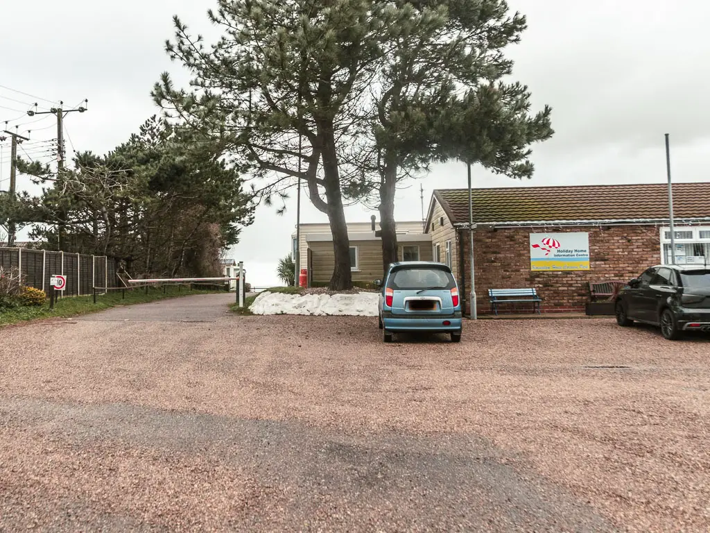 A car parking area with a small single story budding ahead. There are two parked cars. There are two tall trees next to the building on the left.