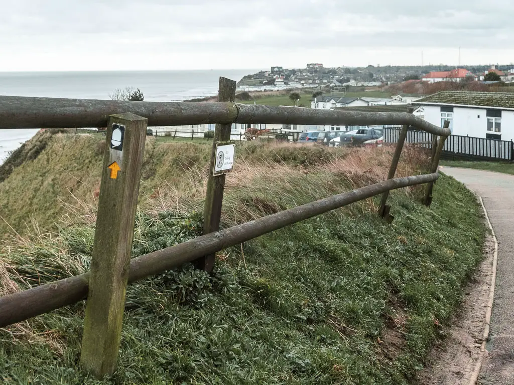 A wooden pole fence curving along the grass bank on the side of the road. There is a wooden sign next to the fence with a white acorn and yellow arrow pointing ahead along the road.