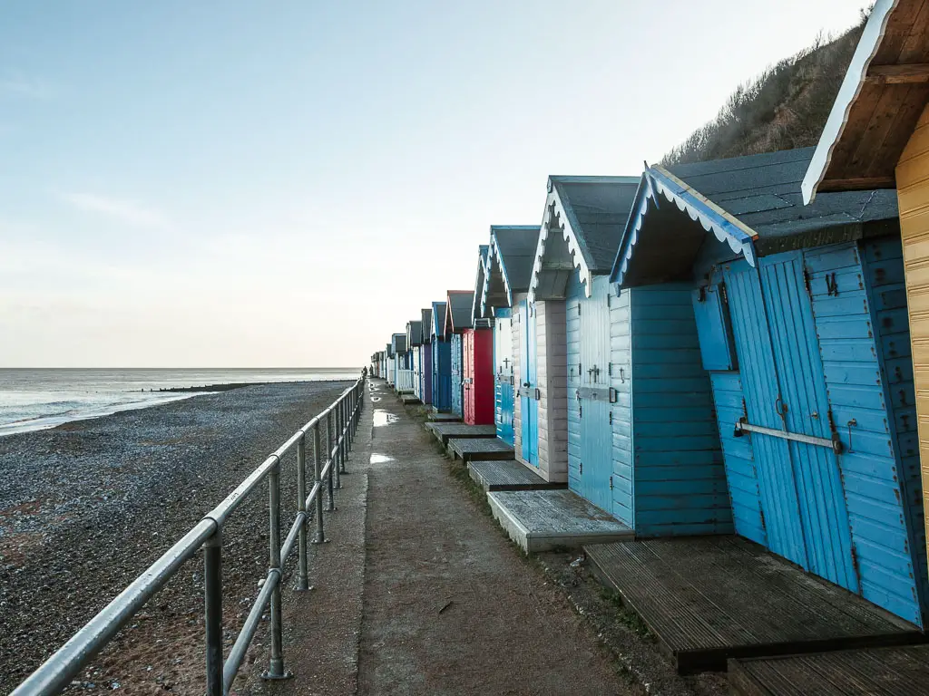 A narrow concrete walkway in Cromer, with colourful beach huts on the right and a black shingle beach to the left, at the start of the walk to Mundesley.