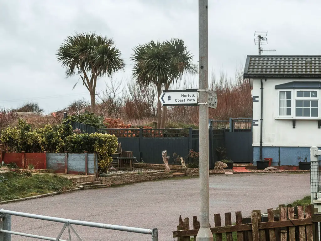 A white arrow sign on a metal pole. The sign points left. There is a driveway behind the sign with the end of a white walled house on the right. 