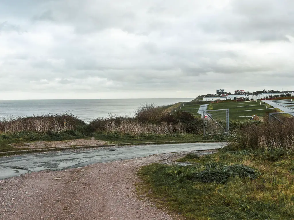 A gravel path leading onto a road, with a view to the grass clifftop and village ahead in the distance. 