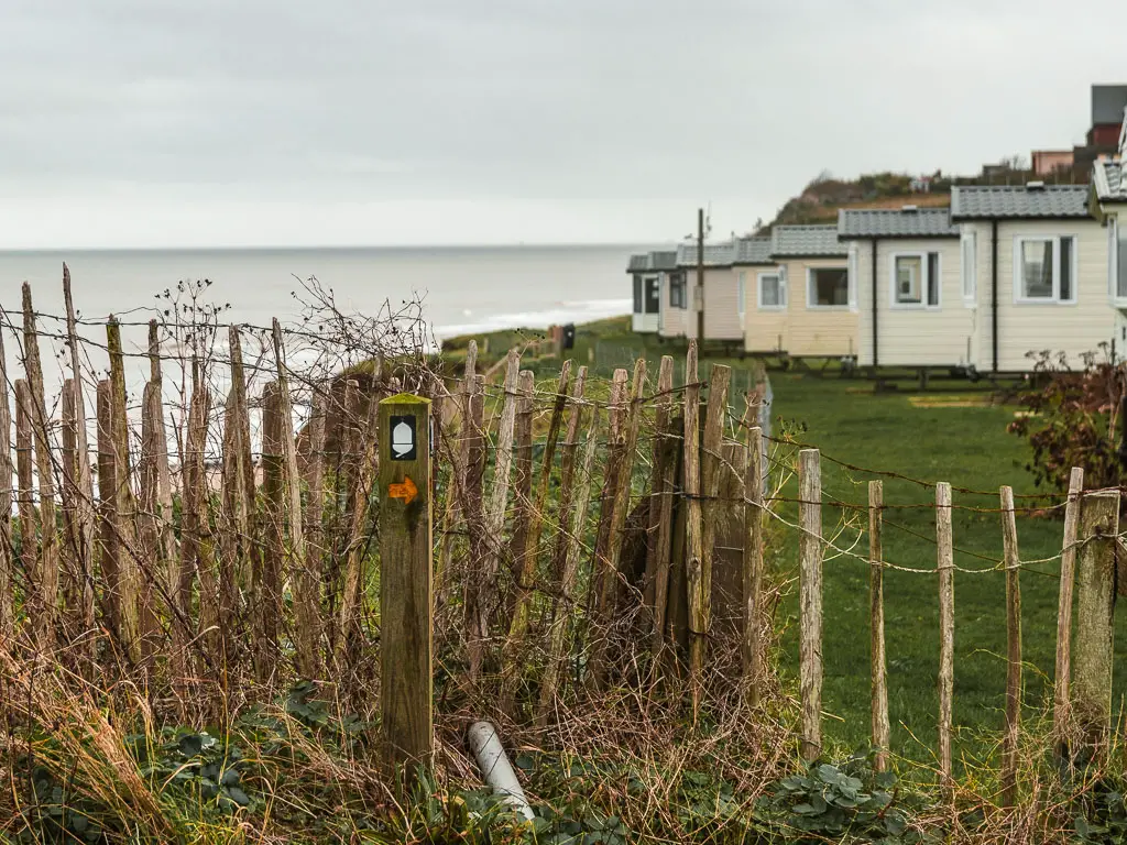 A wooden stump sign with white acorn and arrow pointing right, near the end of the walk from Cromer to Mundesley. There is a rugged wooden fence behind the sign, with a grass area and row of holiday homes on the other side of the fence. 