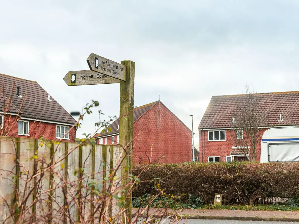 A wooden trail signpost on a fence corning next to the road, with red bricked houses on the other side of the road.