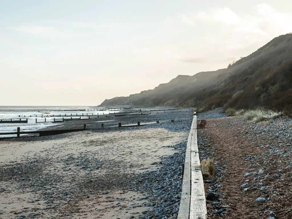 Looking along a long stretch of beach, with sand and black shingle on the left, and dirt and black rocks on the right, with a wooden wall in between. there are rugged cliffs looming above to the right.