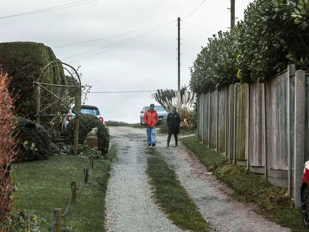 A gravel driveway style road lined with a grass patch on the left and tall wooden fence on the right. There are tow people walking down the road. 