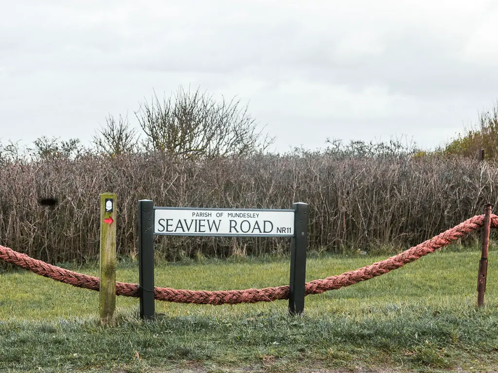 A road sign saying 'Seaview road parish of Mundesley'. the sign is sitting low on the grass, with a red rope hanging behind it. There us a trail sign with white acorn, and red arrow pointing right.