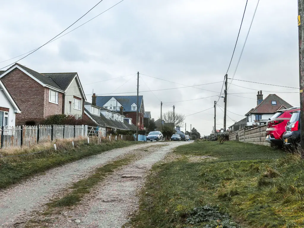 A driveway road lined with houses on the left, and cars parked on the right and ahead on the left. There are telephone pylon wires above.