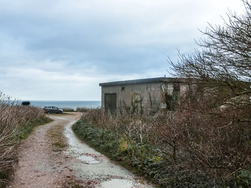 A road curving ahead, lined with leafless bushes, and a shelter on the right. The blue sea is visible in the distance ahead. 