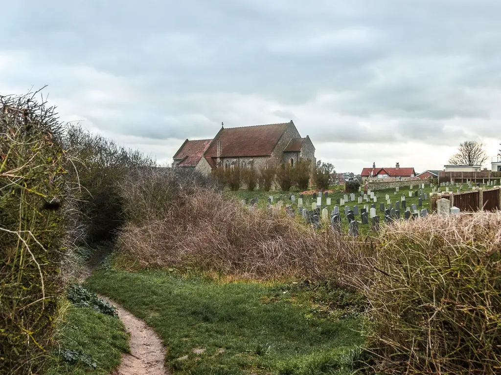 A thin dirt trail curving to the left through the bushes, with a graveyard and church on the right, at the end of the walk from Cromer to Mundesley.