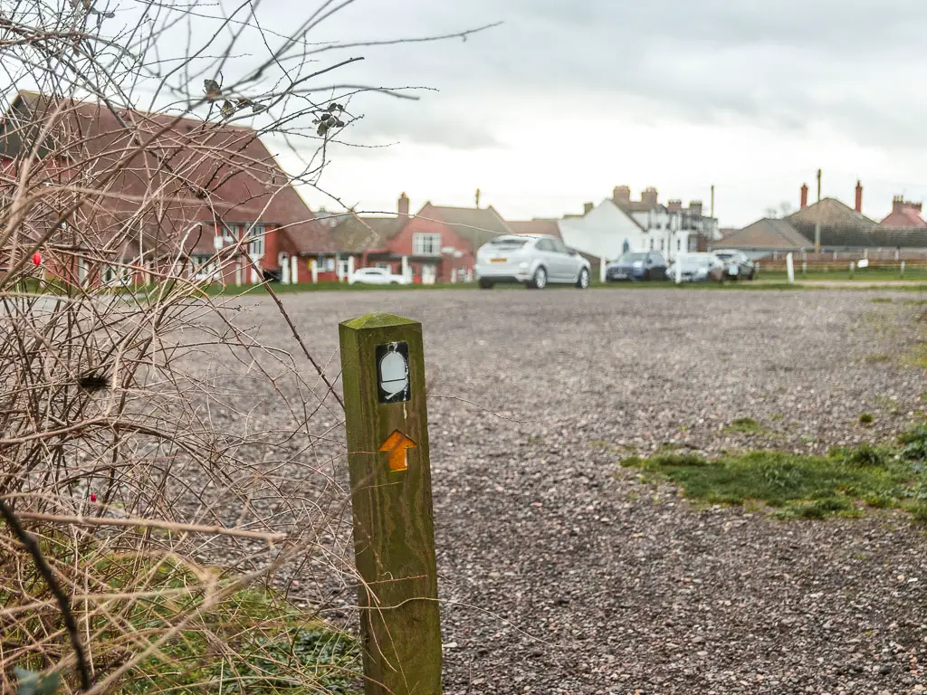 A wooden stump trail sign with white acorn and yellow arrow pointing ahead across the large gravel car park. There are a few cars parked on the other side, with houses behind them.