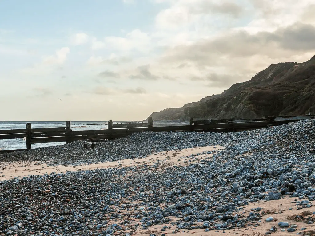 Looking along a beach covered in grey small rocks, with patches of sand, and cliffs on the left, on the walk between Cromer and Mundesley.