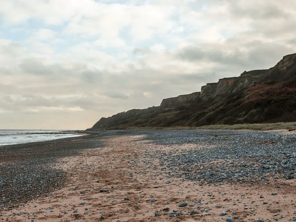 Looking along a long stretch of beach covered in grey shingle with patches of sand, on the walk from Cromer to Mundesley. There are rugged cliffs over on the right.