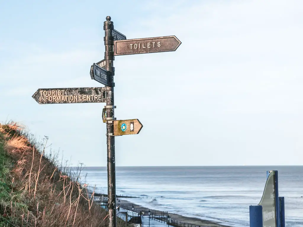 A metal sign with arrows pointing in all directions, at the start of the walk towards Mundesley from Cromer. The blue sea is visible ahead below.