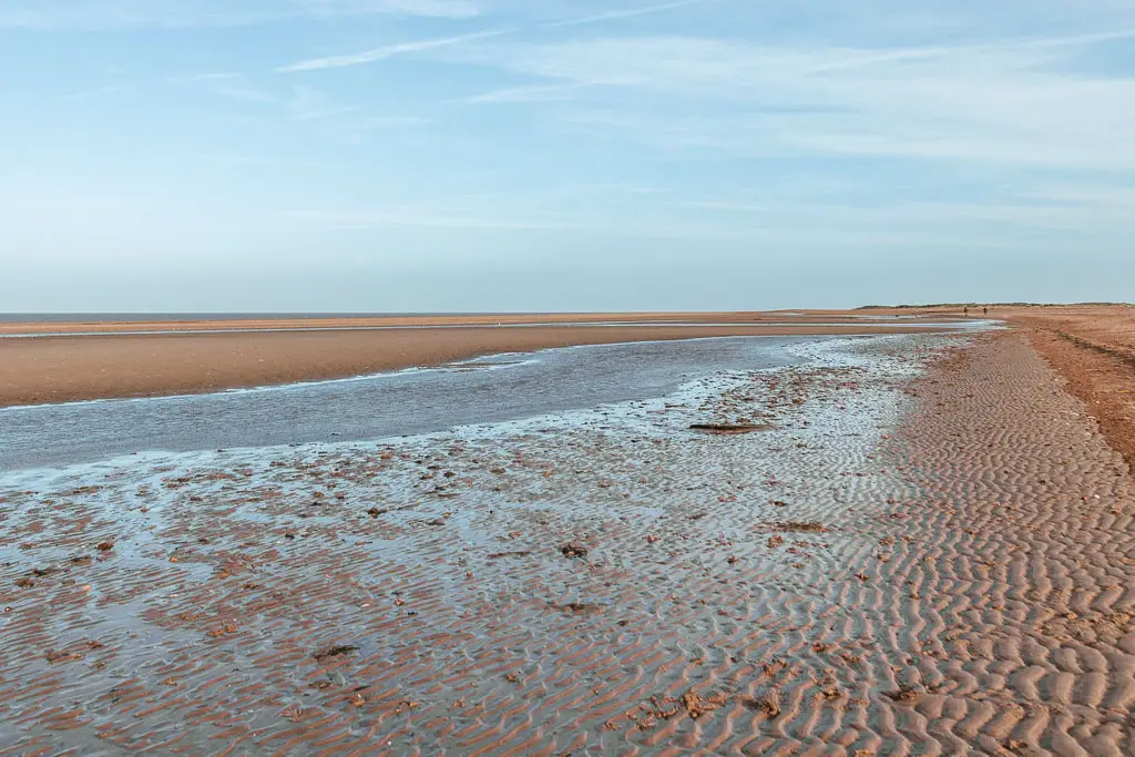 A long stream of water running along the long stretch of sandy beach. The sky is light blue.