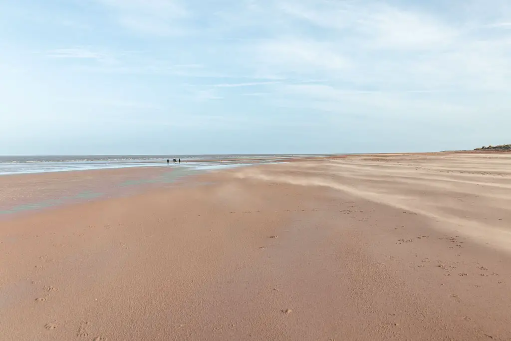 Looking along a very long wide stretch of light beige coloured sand at the start of the walk from. Hunstanton to Burnham Overy Staithe. There are tiny specs of three people walking in the distance. The sky is light blue.