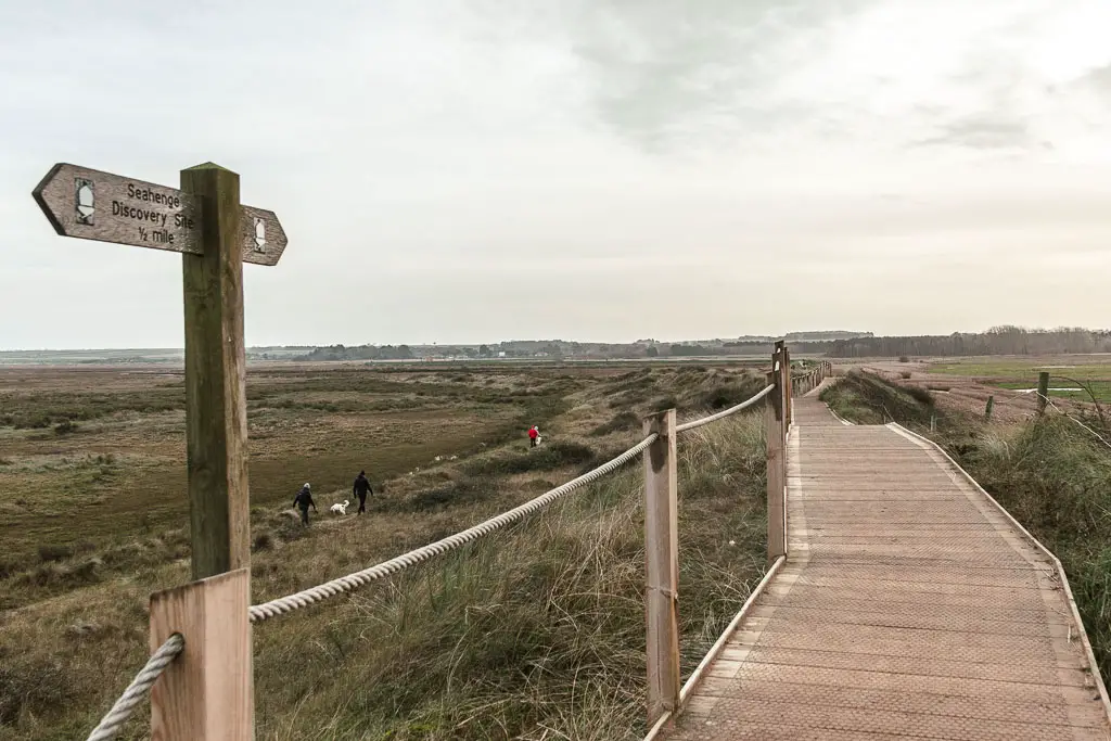 A wooden walkway, with a field of grass and marsh to the left. There is a trail signpost on the left side of the walkway, and a roped fence. 