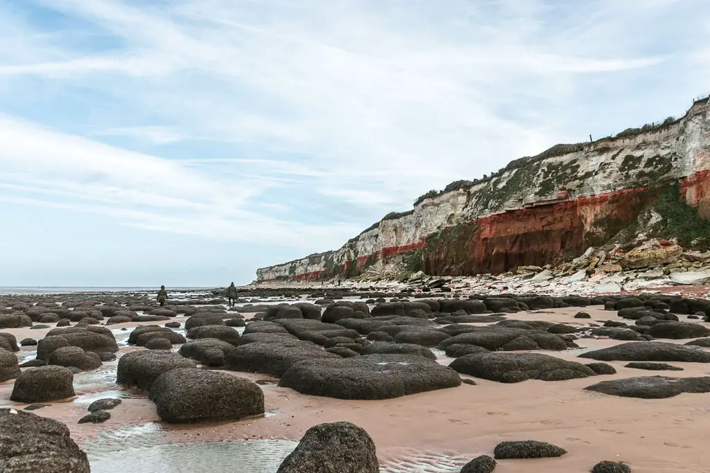 A sandy beach with black boulders scattered about, and the red and white stripped Hunstanton cliffs on the other side, on the walk towards Burnham Overy Staithe. There are two people walking across the boulders ahead. 